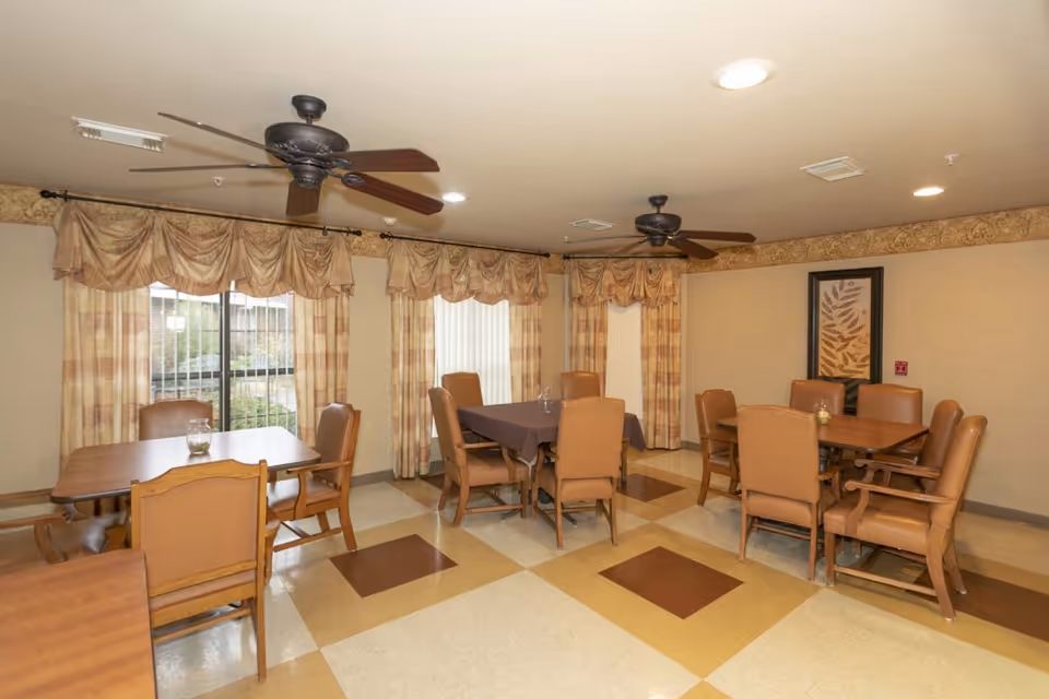 A dining room with multiple wooden tables and tan cushioned chairs arranged around them. The room has large windows with patterned curtains, two ceiling fans with lights, and a framed artwork on the wall. The floor has a checkered pattern with beige and brown tiles.