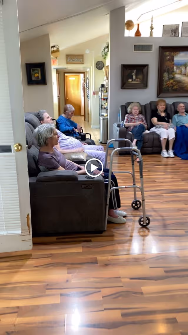 Elderly residents seated in recliners in a senior home common room with a walker and wood flooring.