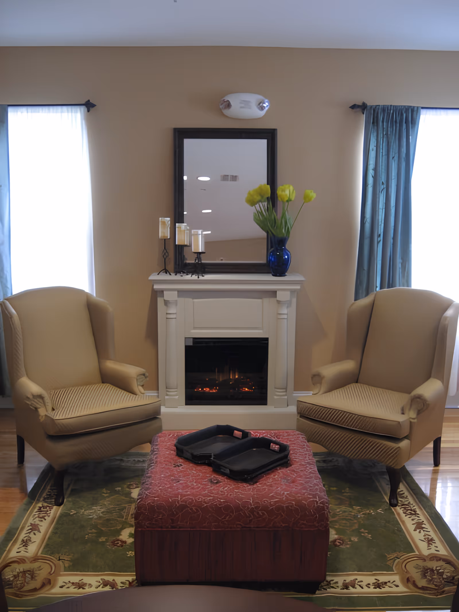 A cozy living room area with two beige armchairs facing each other, a red patterned ottoman in the center with two black trays on top, a white fireplace with a mirror above it, and a blue vase with yellow flowers on the mantel. The room has light brown walls and two windows with blue curtains on either side of the fireplace.
