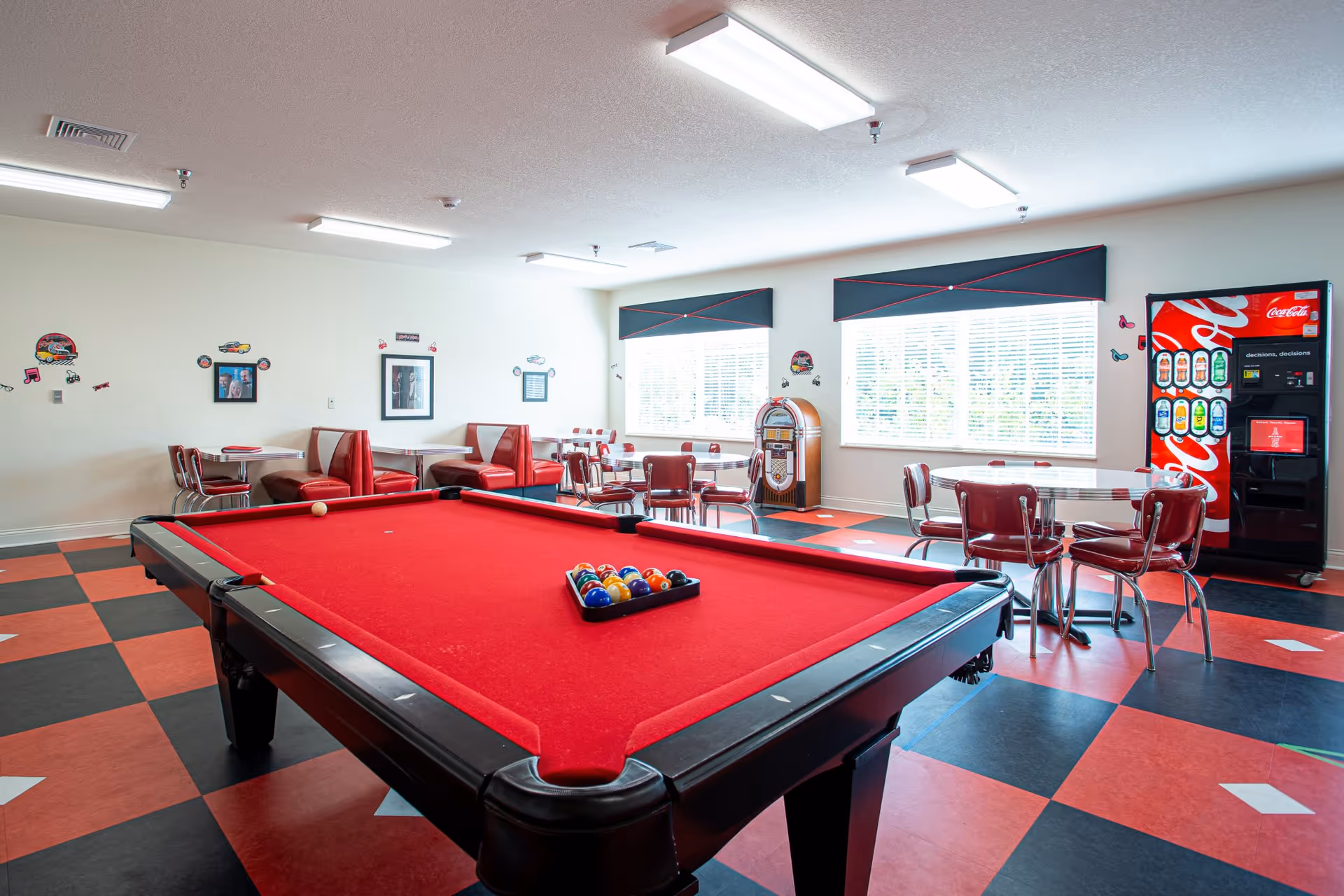 A recreational room with a red felt pool table in the foreground, surrounded by retro-style red and white booths and chairs. The room features a black and red checkered floor, large windows with blinds, a vintage jukebox, and a Coca-Cola vending machine. The walls are decorated with small car and music-themed decals.