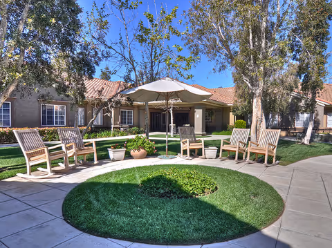 Outdoor seating area at a senior living facility with wooden rocking chairs arranged in a circle around a large umbrella. The area is surrounded by green grass, trees, and a building with a tiled roof and multiple windows in the background under a clear blue sky.