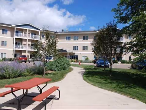 Exterior view of Pine Manor Estates with a path, trees, lawn, and a red picnic table.