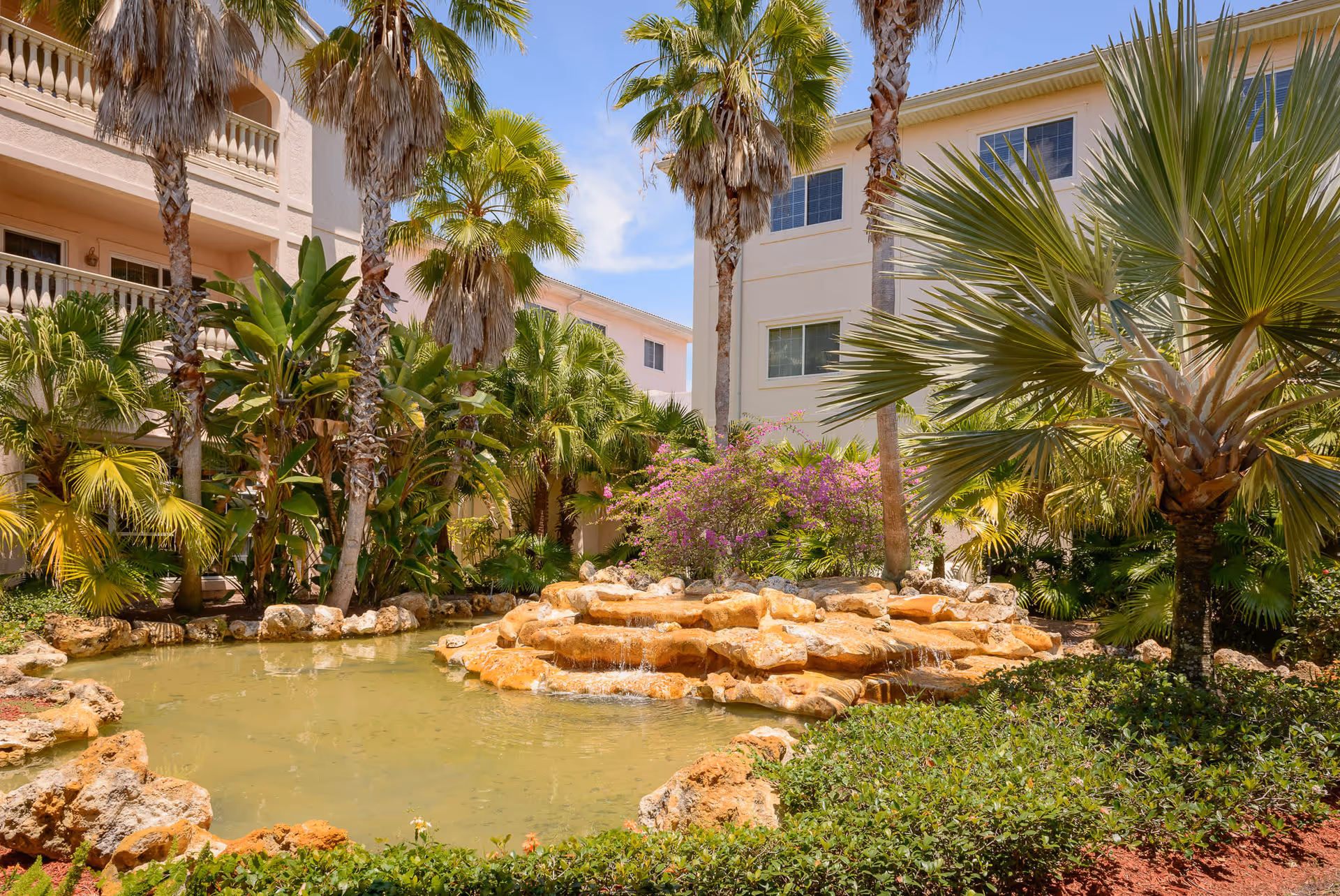 A landscaped outdoor area with a small pond and rock waterfall surrounded by lush tropical plants and palm trees, with light-colored residential buildings in the background under a blue sky.