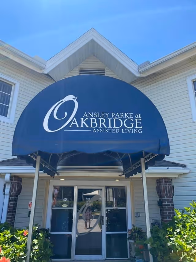 Entrance of Ansley Parke at Oakbridge Assisted Living facility with a blue awning displaying the facility's name above glass double doors, surrounded by light-colored siding and some greenery.