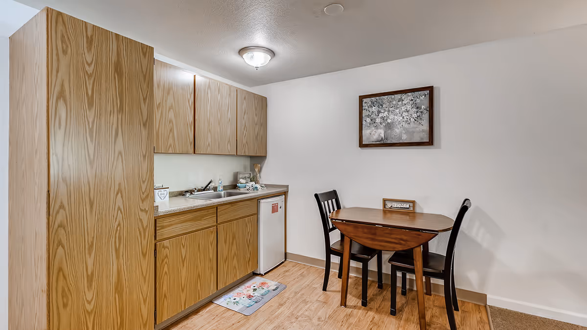A small kitchen area with wooden cabinets, a countertop with a sink, and a mini refrigerator. Next to the kitchen is a wooden drop-leaf table with two black chairs. A framed floral painting hangs on the white wall above the table, and a small decorative sign that says 'HOME' is on the table.