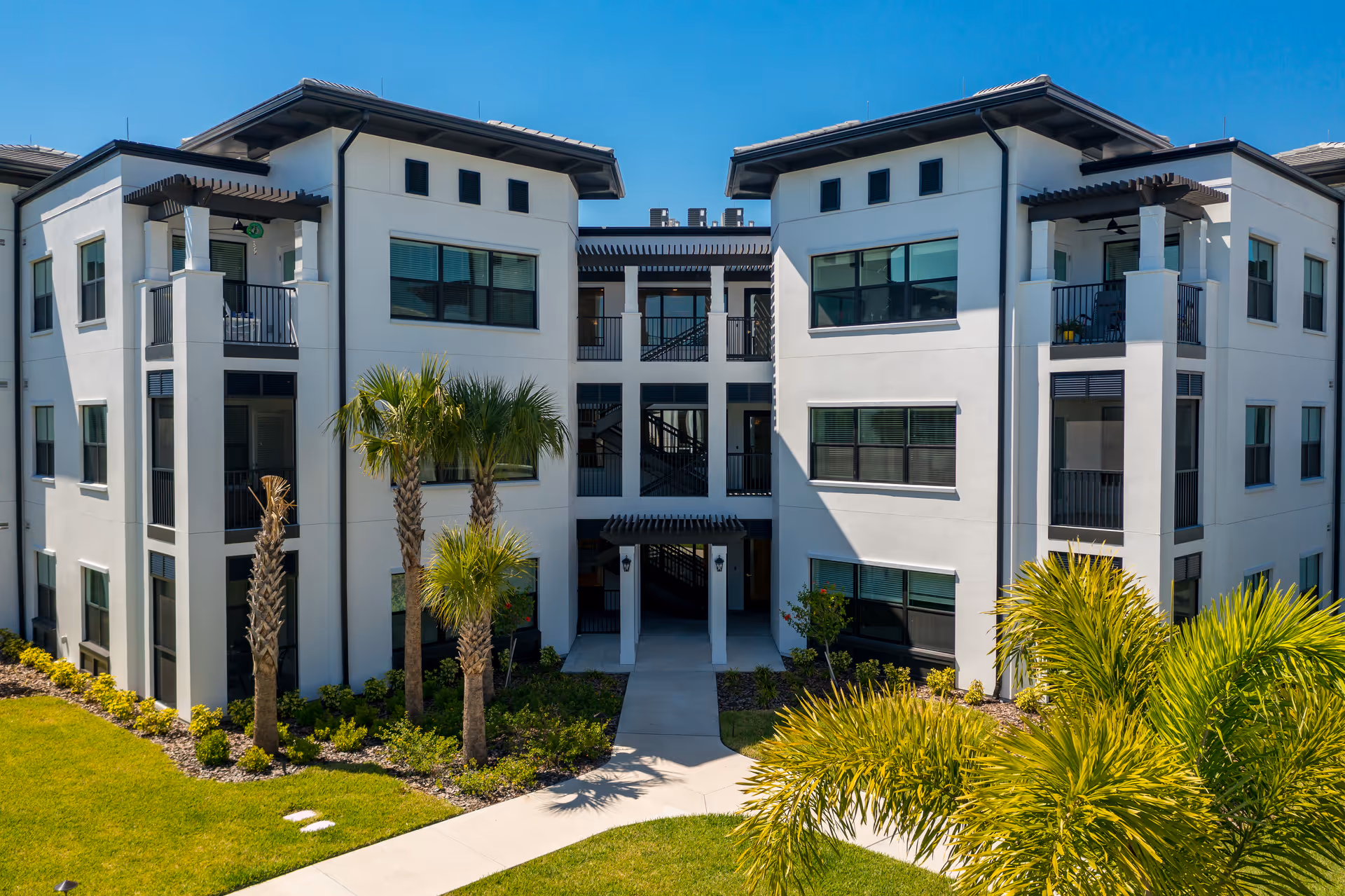 Exterior view of a modern three-story residential building with white walls and large windows, surrounded by palm trees and landscaped greenery under a clear blue sky.