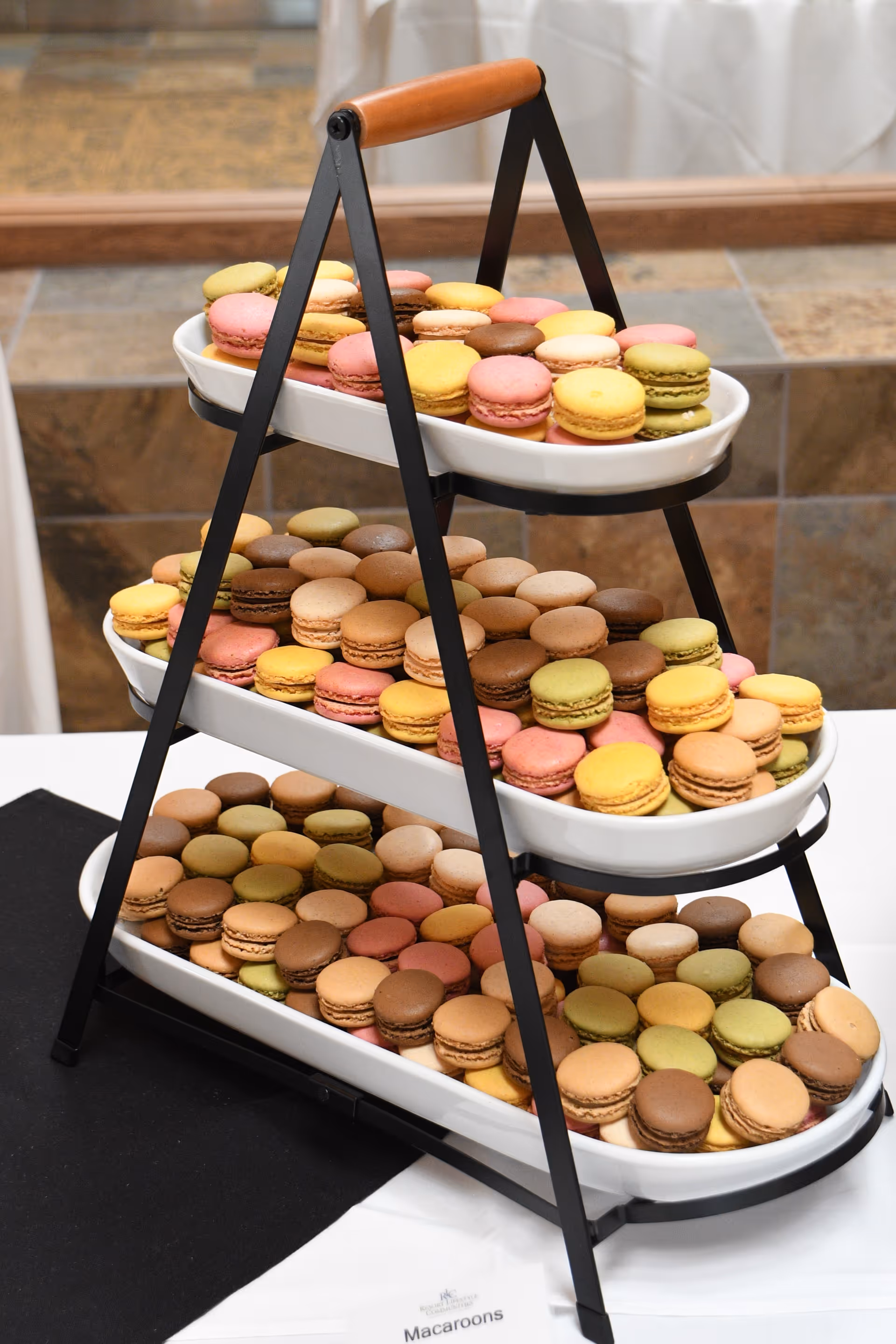 Three-tiered serving tray filled with colorful macarons in various flavors and colors, placed on a table with a white tablecloth and a black placemat, with a tiled wall in the background.