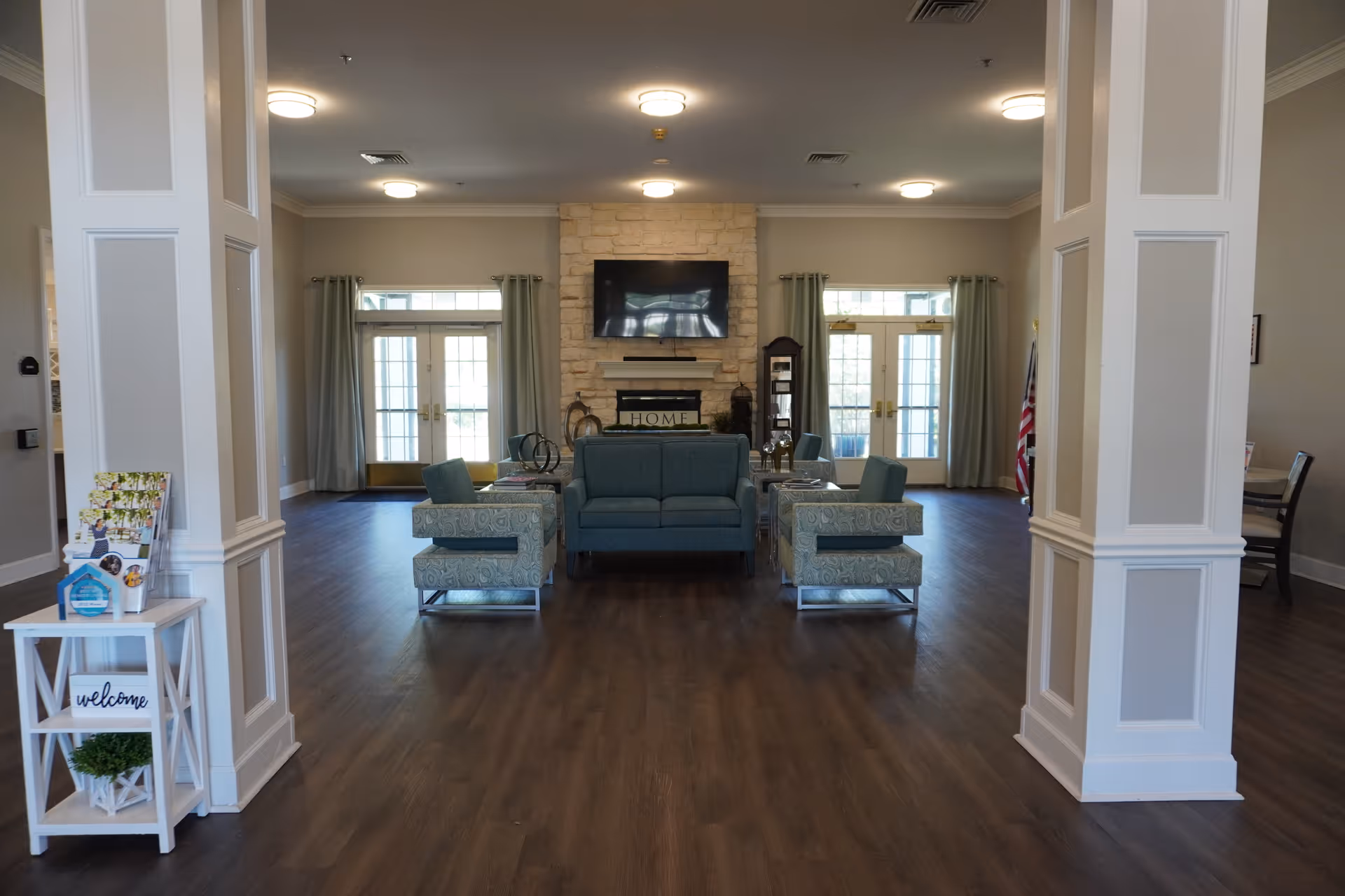 A spacious living room area in a senior living facility with a central seating arrangement including a blue sofa and four patterned armchairs. The room features a stone fireplace with a mounted flat-screen TV above it, two sets of French doors with curtains on either side, and wooden flooring. There are two white columns framing the seating area and a small white table with a welcome sign and brochures on the left side.