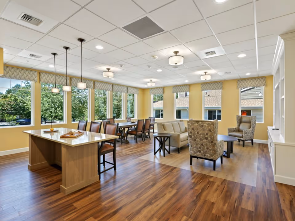 A bright and spacious common area with large windows letting in natural light. The room features a wooden floor, a central island with chairs, a dining table with chairs, and a seating area with a sofa and patterned armchairs. The walls are painted yellow and the ceiling has recessed lighting and hanging pendant lights.