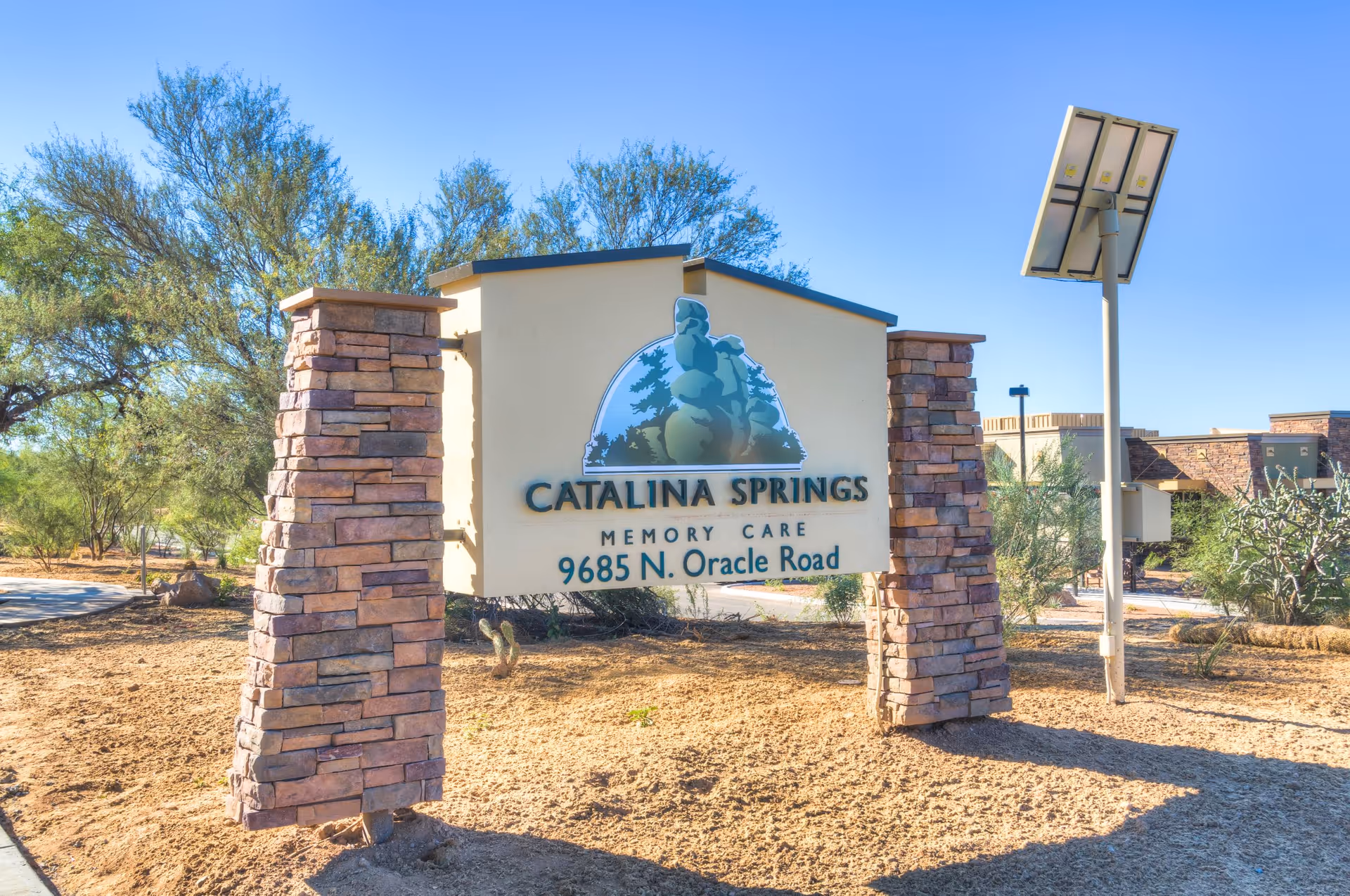 Outdoor sign for Catalina Springs Memory Care located at 9685 N. Oracle Road, supported by two stone pillars with desert vegetation and a solar panel nearby under a clear blue sky.