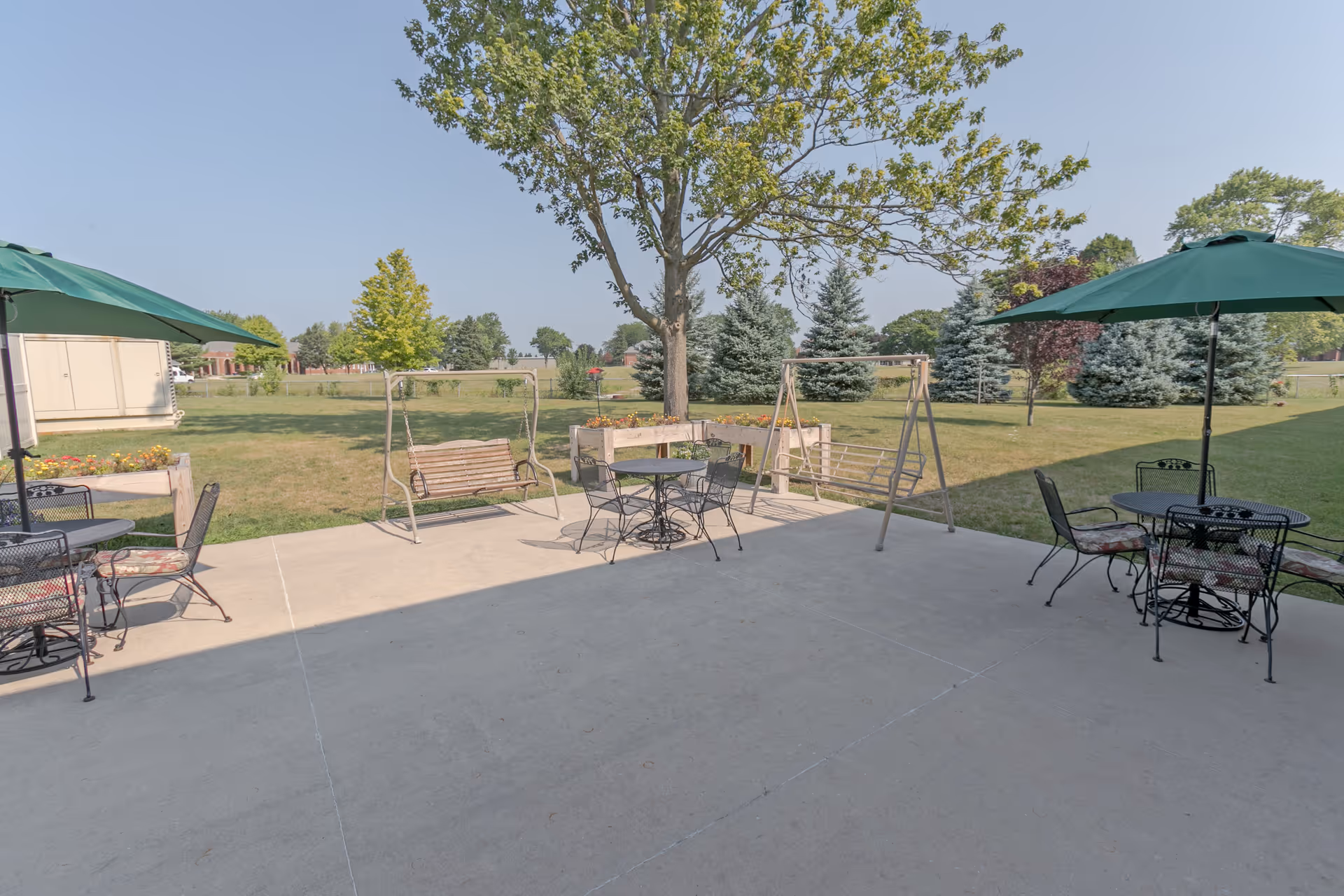 Outdoor patio with tables, umbrellas, and swing benches overlooking a grassy yard and trees.