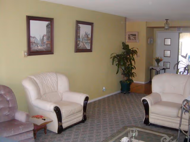 A living room area with two white armchairs and one purple armchair arranged around a glass coffee table. The walls are painted light green and decorated with framed pictures. There is a potted plant near the back wall and a small table with flowers near an exit door in the background.