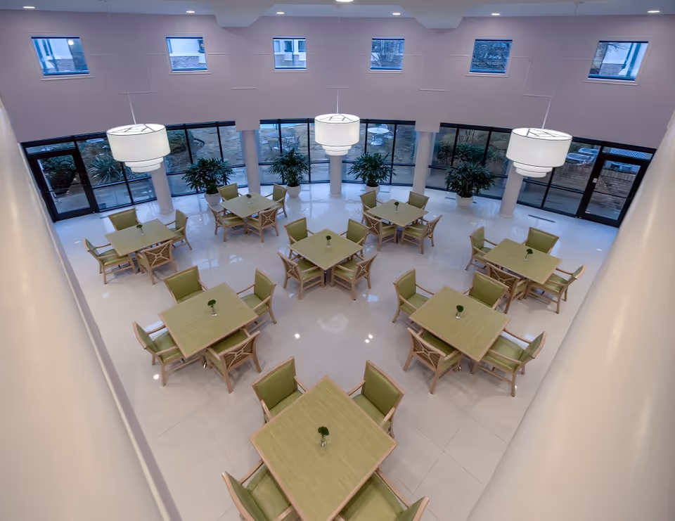 Bright atrium dining room with multiple square tables and green chairs arranged on a glossy tiled floor beneath large pendant lights and tall windows.