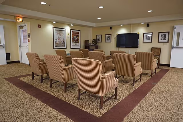 A small theater-style room with two rows of striped armchairs facing a wall-mounted flat screen TV. The walls are decorated with framed pictures and movie posters. The room has beige walls, recessed lighting, and a patterned carpet with a maroon border.