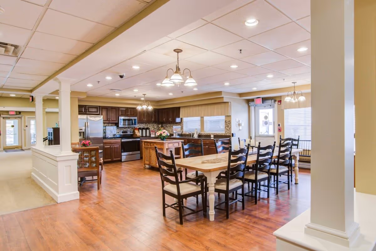 A spacious dining area with a long wooden table surrounded by black chairs with cushioned seats. The room features a kitchen in the background with dark wood cabinets, stainless steel appliances including a refrigerator, oven, and microwave. The floor is wooden, and the ceiling has recessed lighting and hanging light fixtures. Large windows with blinds allow natural light into the room.