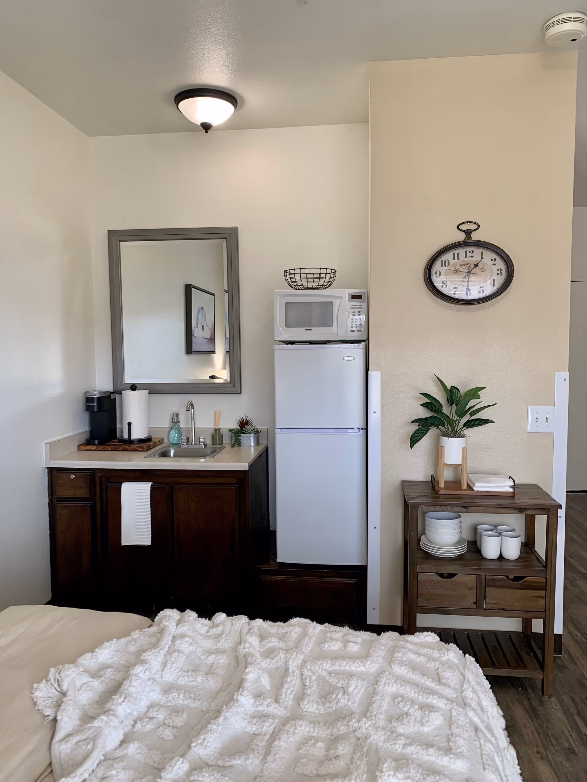 A small kitchenette area in a senior living facility room featuring a dark wood cabinet with a sink, a coffee maker, and a paper towel holder. Above the cabinet is a rectangular mirror. Next to the cabinet is a white refrigerator with a microwave on top and a wire basket. To the right, there is a wooden side table with a potted plant, stacked plates, and cups. A round wall clock hangs above the side table. The foreground shows part of a bed with a textured white blanket.