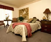 Cozy furnished bedroom with a bed, nightstand and lamp, seating, window with curtains, and wall art.