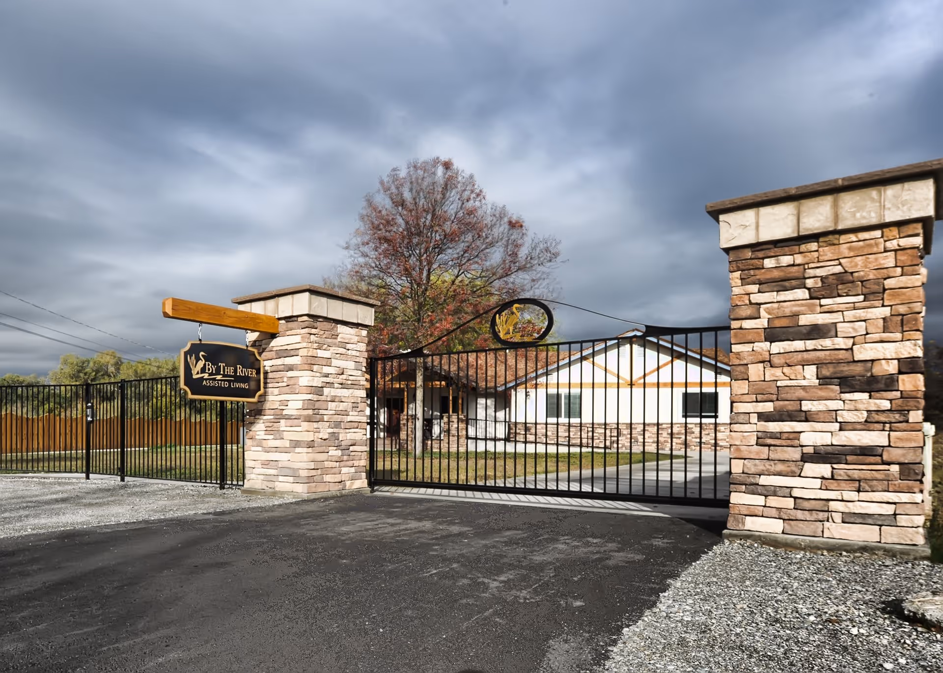 Entrance gate to By the River Assisted Living facility with stone pillars and a black metal gate, a sign hanging on the left pillar, and a building with white walls and stone accents in the background under a cloudy sky.