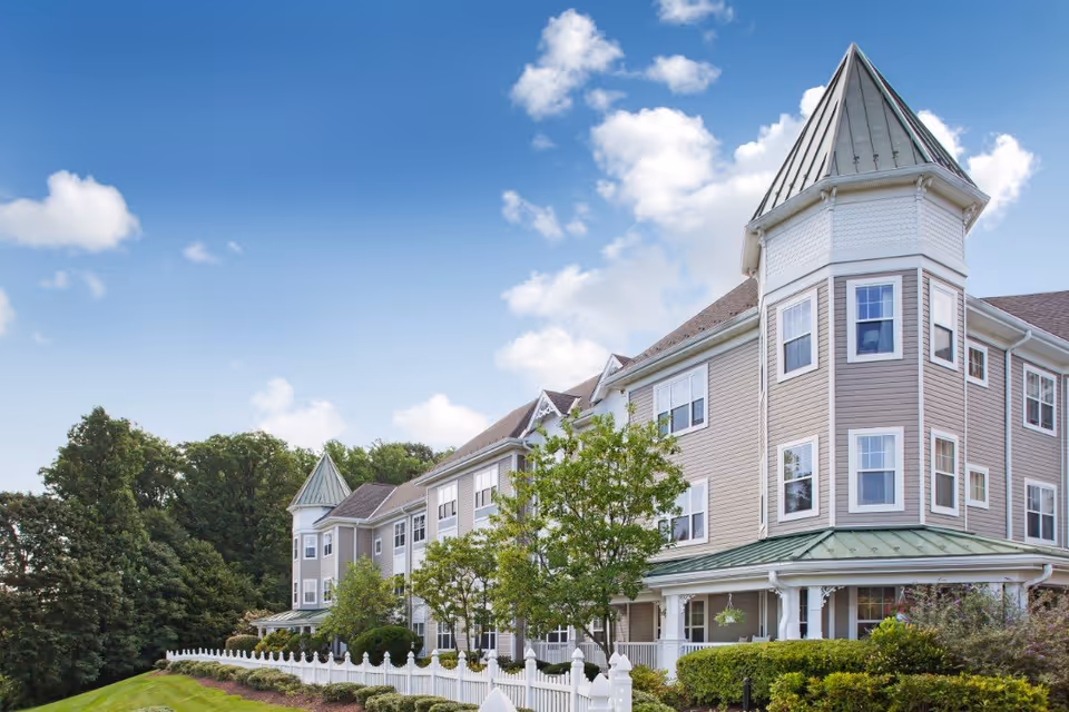Exterior view of a large, multi-story senior living facility with beige siding, white trim, and a green metal roof. The building features a prominent turret and multiple windows. There is a white picket fence and well-maintained landscaping with trees and bushes in front of the building under a blue sky with scattered clouds.