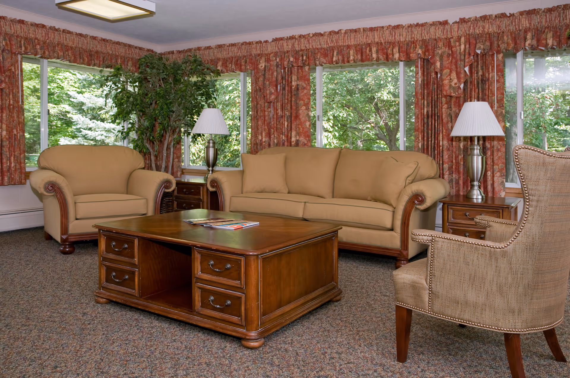 Sunlit living room with beige sofa and armchairs, a wooden coffee table, side tables with lamps, and large windows with patterned curtains.