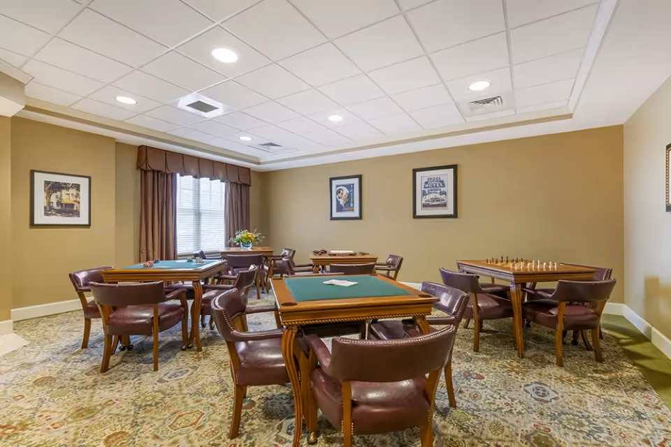 A well-lit game room with multiple card tables and leather chairs arranged around them. The room has beige walls, a patterned carpet, framed artwork on the walls, and a window with brown curtains letting in natural light.