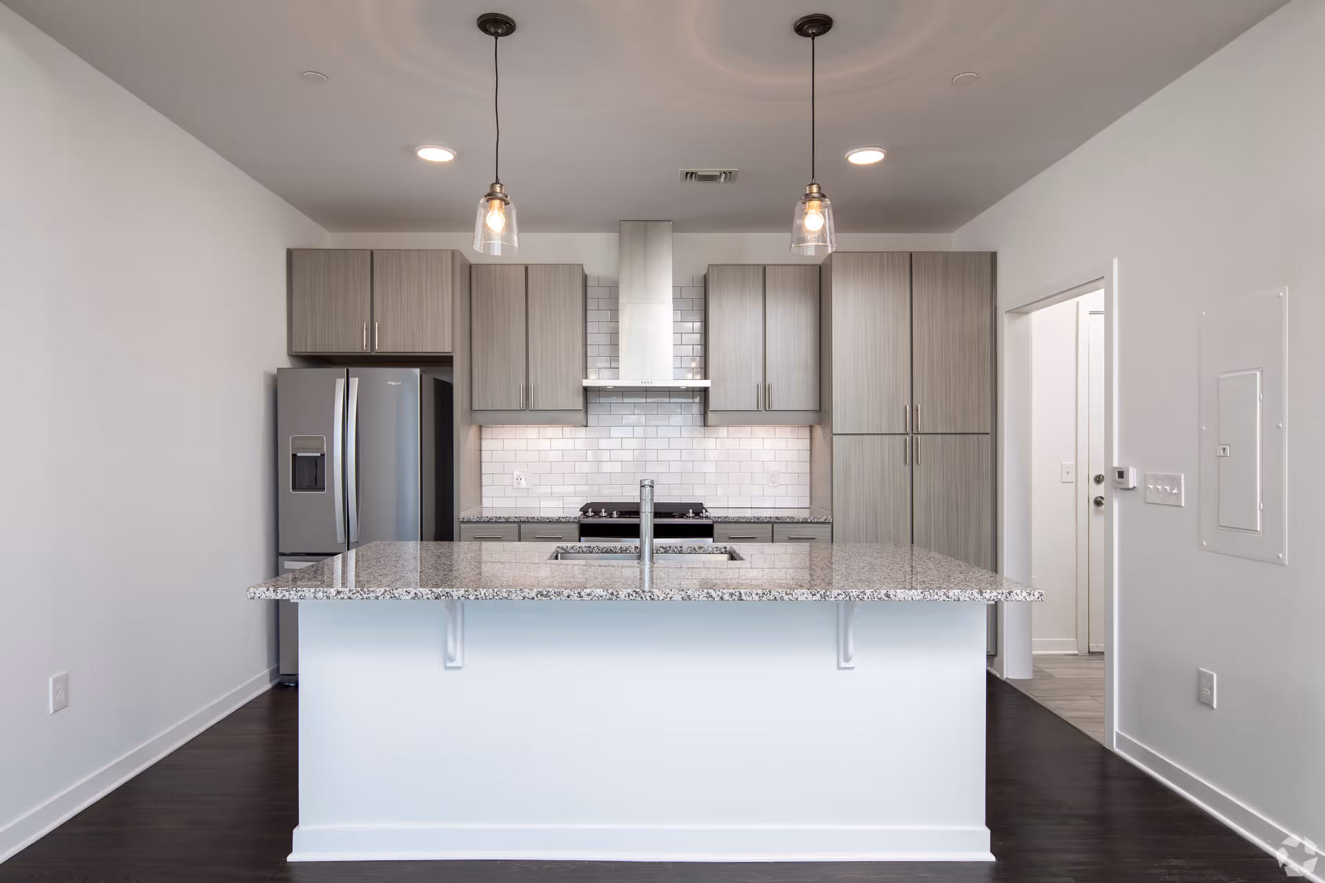 Modern kitchen with a large granite island countertop, stainless steel refrigerator, stove with a stainless steel hood, gray cabinets, white subway tile backsplash, and two pendant lights hanging from the ceiling.