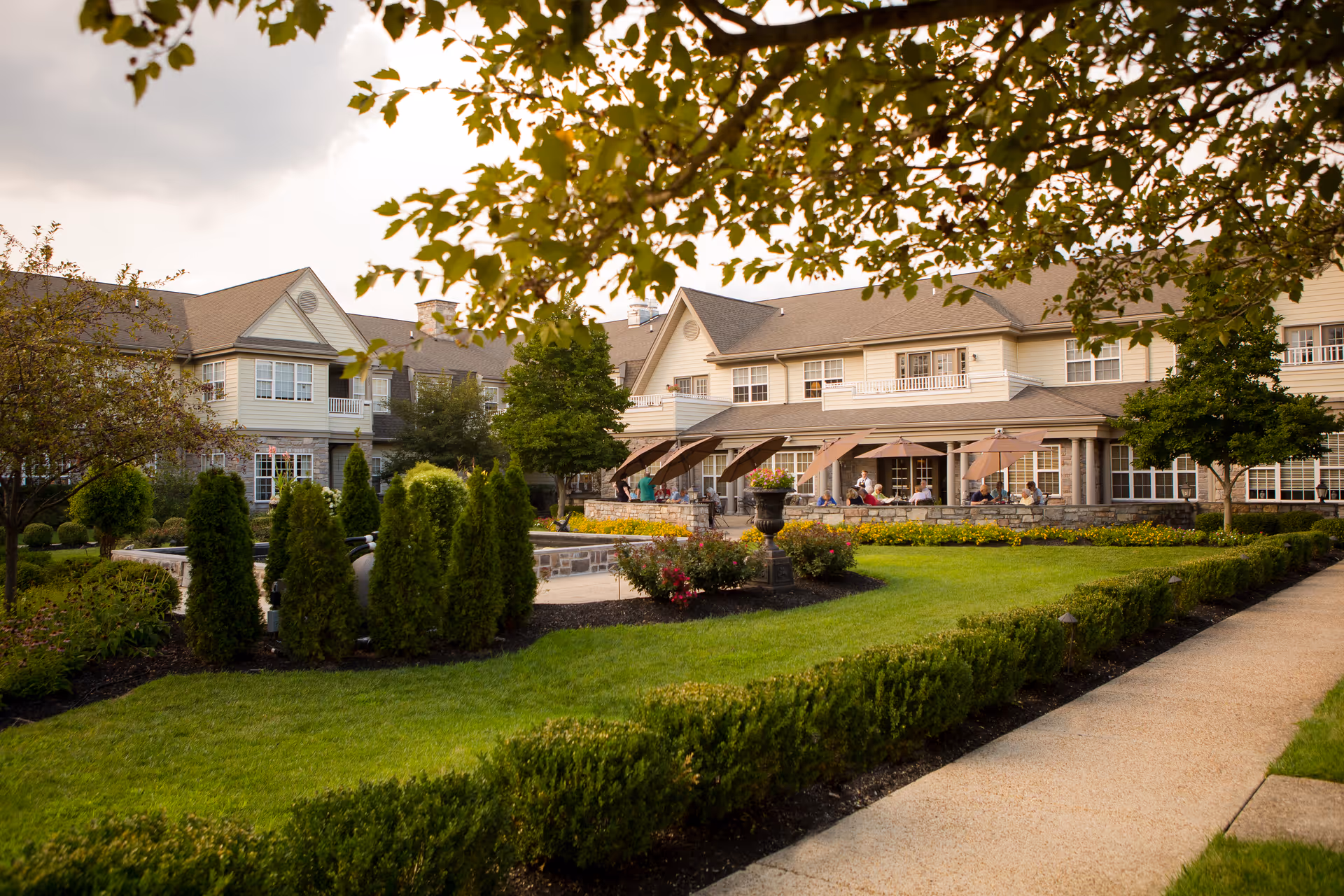 Front exterior of a senior living building with landscaped lawns, terrace seating, and a paved walkway.