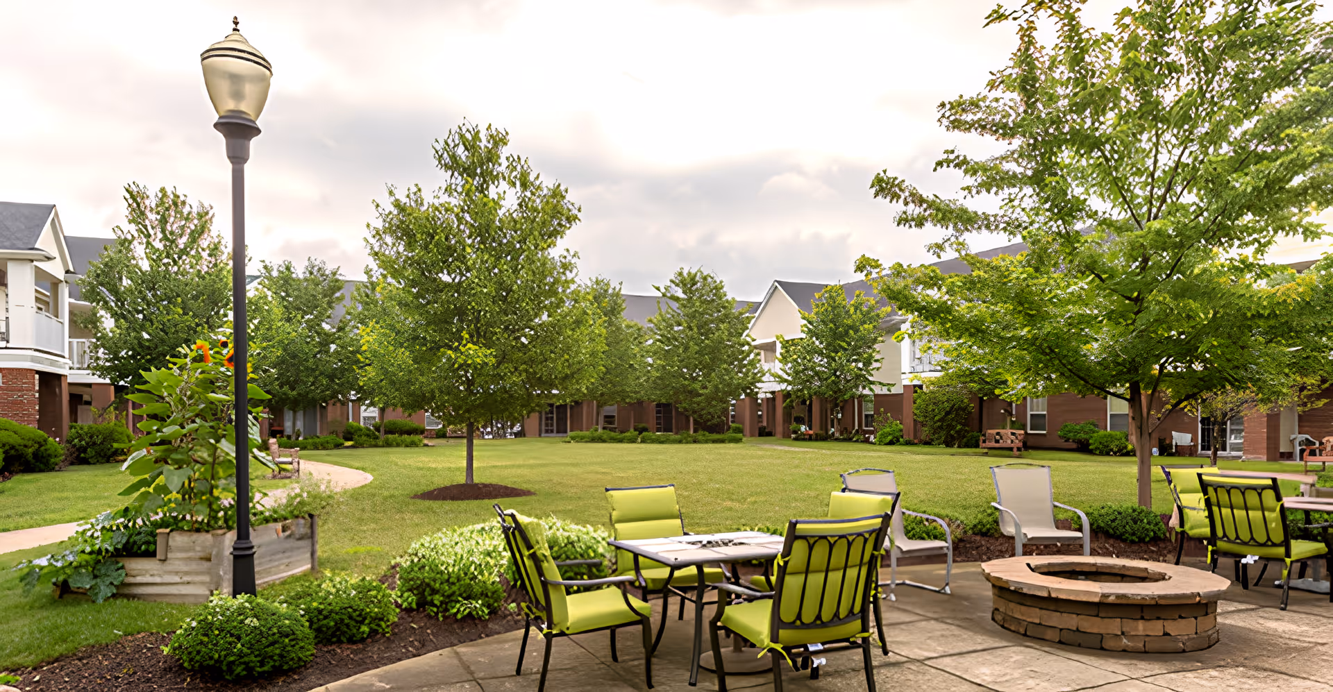 Outdoor patio area at Independence Village of Aurora featuring green cushioned chairs around a fire pit and tables, surrounded by well-maintained grass, trees, and shrubs with residential buildings in the background under a cloudy sky.