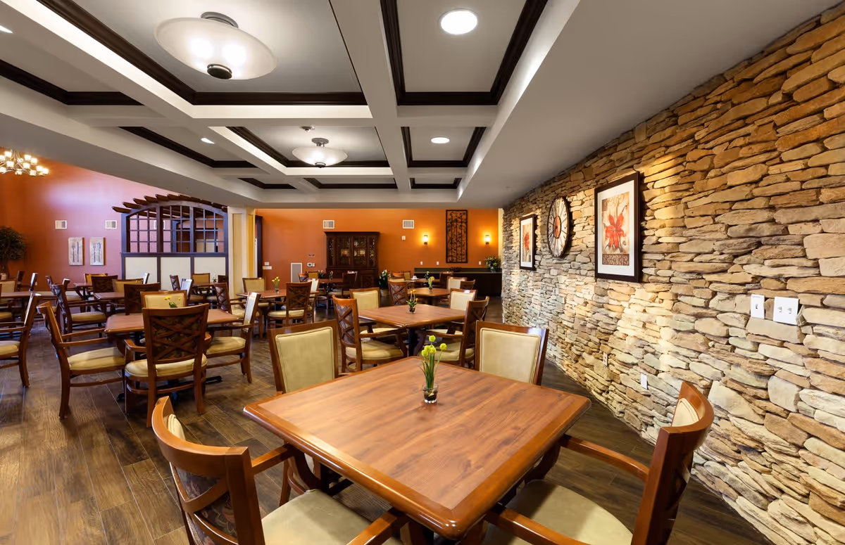 A spacious dining room with wooden tables and cushioned chairs arranged neatly. The room features a stone accent wall with framed artwork and a large clock. The ceiling has a coffered design with recessed lighting and modern light fixtures. The floor is wooden, and small flower vases are placed on the tables.