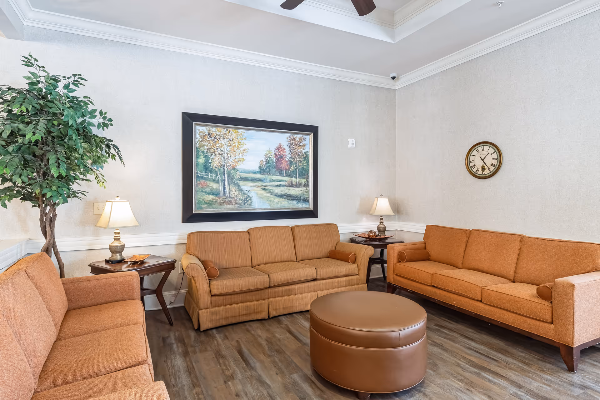 Sitting room with three orange sofas arranged around a round leather ottoman, side tables with lamps, a large landscape painting, a wall clock, and a potted plant.