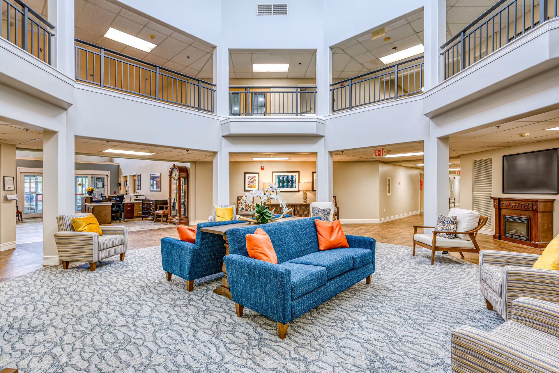 A spacious and well-lit senior living facility common area with blue sofas adorned with orange pillows, striped armchairs with yellow pillows, a patterned carpet, and a wooden fireplace with a mounted TV. The area features a two-story ceiling with a balcony railing on the upper floor and several doorways leading to other rooms.