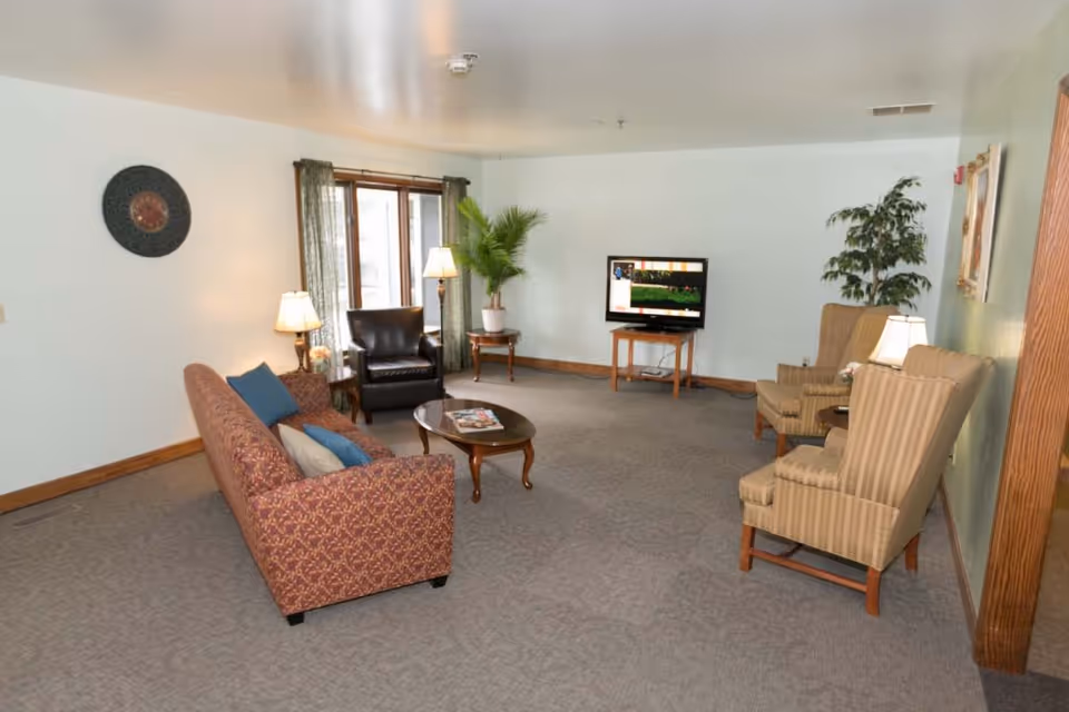 A living room in an assisted living facility with a patterned sofa, two striped armchairs, a black leather chair, a wooden coffee table with magazines, a TV on a wooden stand, two table lamps, green curtains, and potted plants.