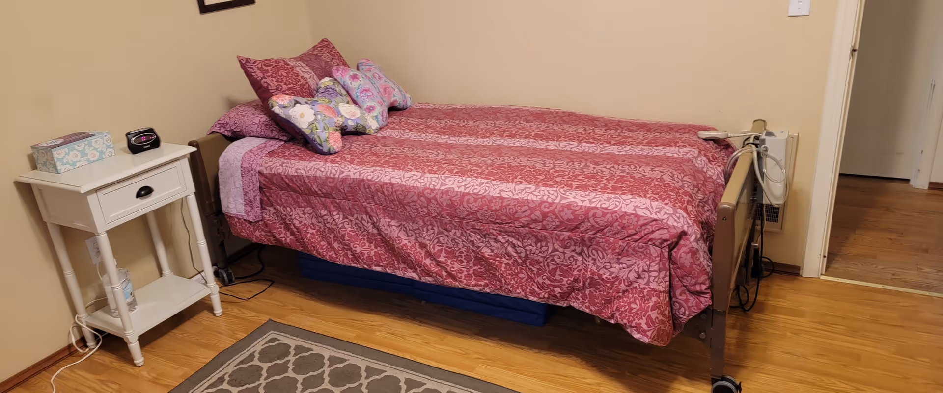 A single bed with a pink and purple patterned bedspread and multiple decorative pillows in a small room. Next to the bed is a white nightstand with a tissue box, a clock, and a bottle of hand sanitizer on the lower shelf. The floor is wooden with a patterned rug partially visible. The bed has wheels and medical equipment attached to the headboard.