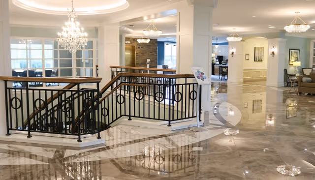 Spacious and elegant interior of a senior living facility featuring a polished marble floor, a black metal railing staircase with wooden handrails, multiple chandeliers, and comfortable seating areas with sofas and lamps. The area is well-lit with natural light coming through large windows and has a welcoming, upscale ambiance.