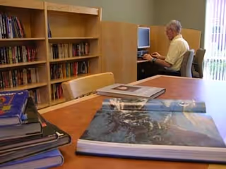 A senior man using a computer in a small library or study area with bookshelves filled with books. In the foreground, there is a wooden table with several large books and photo albums stacked on it. The room has a calm and quiet atmosphere with natural light coming through a window with vertical blinds.