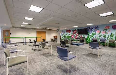 A spacious room with multiple chairs arranged in a socially distanced manner on a carpeted floor. The back wall features a colorful mural of large flowers and greenery, with a large flat-screen TV mounted on it. The ceiling has recessed lighting panels, and there are a few tables and a clock on the far wall.