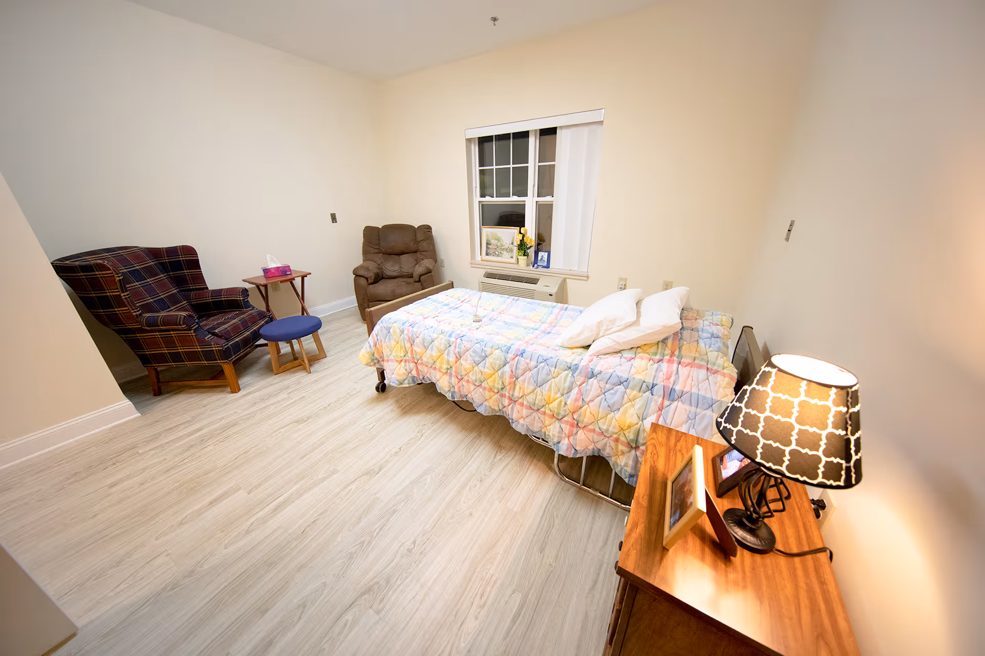A simple bedroom with a single bed covered in a colorful quilt, two pillows, a wooden nightstand with a lamp and framed photos, a window with blinds, a brown recliner chair, a plaid armchair, a small round blue stool, and a small side table with a tissue box. The room has light-colored walls and wood flooring.