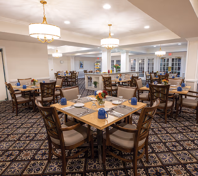 A well-lit dining room with multiple wooden tables and cushioned chairs arranged neatly. Each table is set with blue mugs, plates, silverware, and small floral centerpieces. The room features patterned carpet flooring, white walls, and several elegant ceiling light fixtures. In the background, there are large windows and a small counter area with additional decorations.