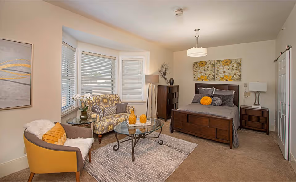 Well-lit bedroom with a bed, a seating area with armchairs and a glass coffee table in front of a bay window, and floral artwork on the wall.