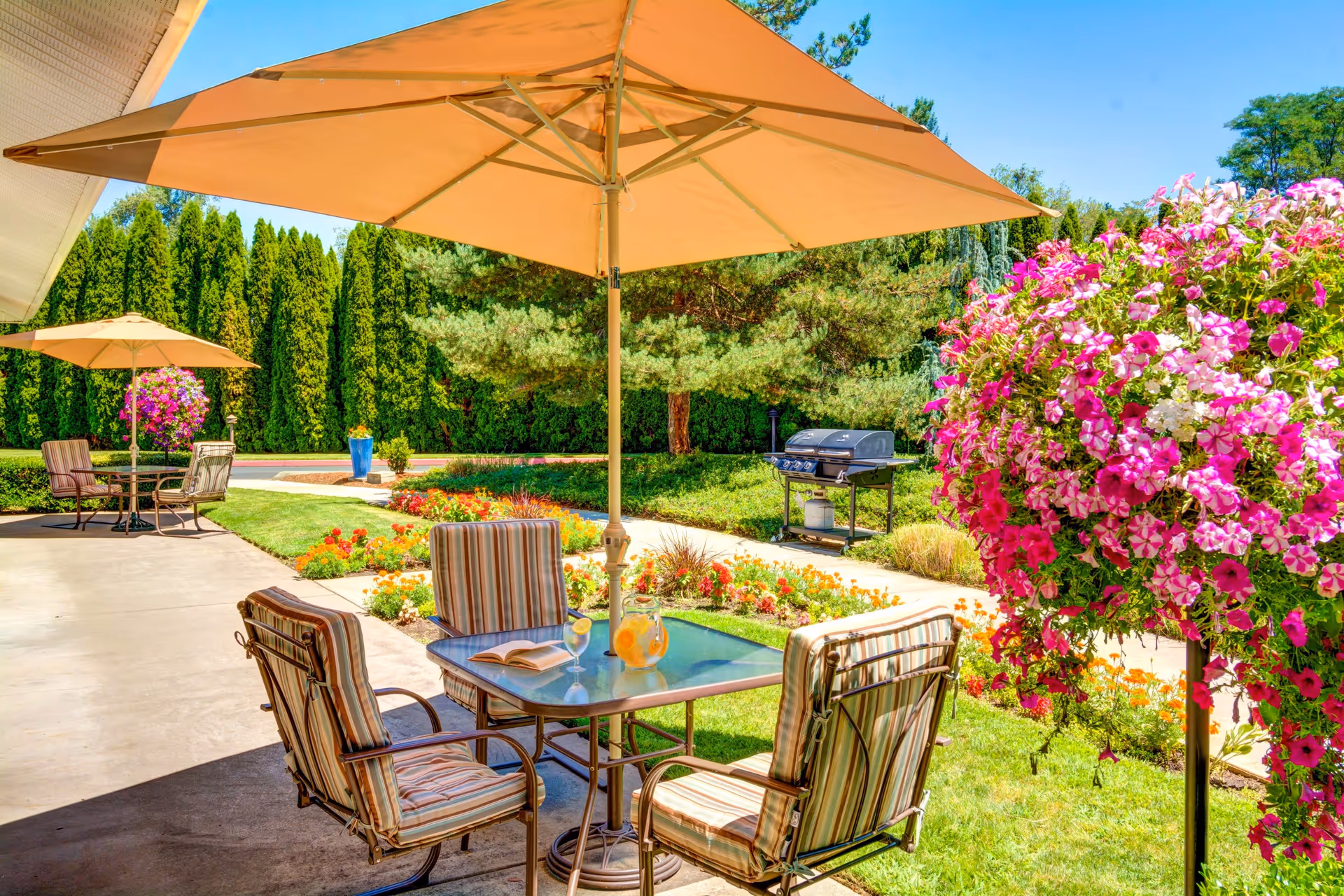 Outdoor patio area with tables and chairs under large beige umbrellas, surrounded by vibrant flowers and greenery. A barbecue grill is visible in the background along a paved walkway, with tall trees and a clear blue sky.