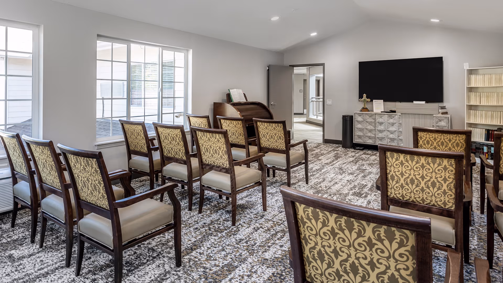 A small meeting or activity room with two rows of wooden chairs with patterned upholstery facing a large flat-screen TV mounted on the wall. The room has a patterned carpet, a roll-top desk near a window, a cabinet under the TV, and bookshelves filled with books. The walls are painted light gray, and there is a door open to a hallway.