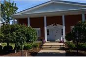 Front entrance of a red-brick building with white columns, double doors, steps and landscaped shrubs.