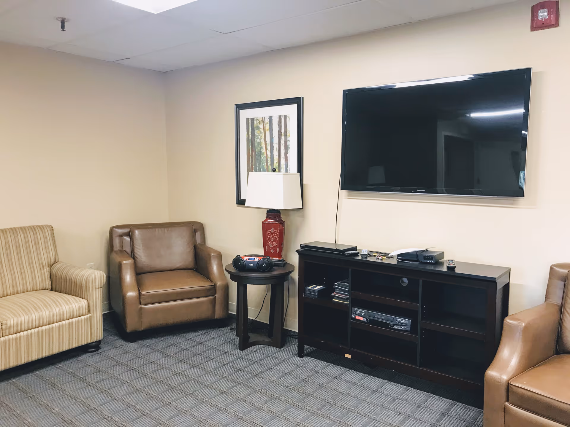 A cozy living room area with a striped beige sofa, two brown leather armchairs, a small round wooden side table with a red lamp and a stereo on it, a wall-mounted flat-screen TV above a dark wooden entertainment unit with various electronic devices and media, and a framed picture of trees on the wall.