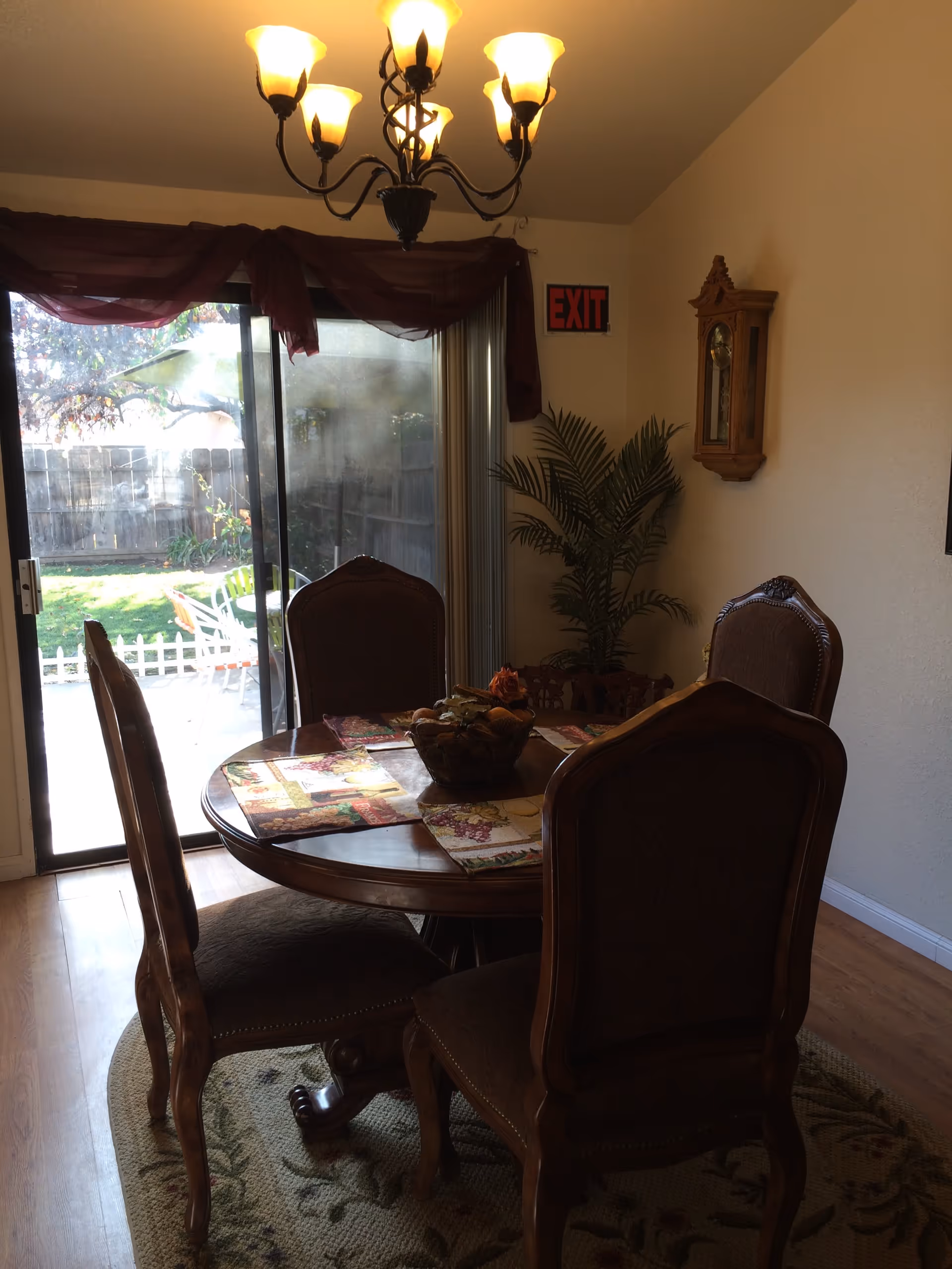 A dining room with a round wooden table surrounded by four upholstered chairs. The table has colorful placemats and a decorative centerpiece. A chandelier with six lights hangs above the table. There is a sliding glass door leading to an outdoor area with a small white fence and garden visible. A potted plant and a wall clock are on the right wall, and an exit sign is mounted above the sliding door.