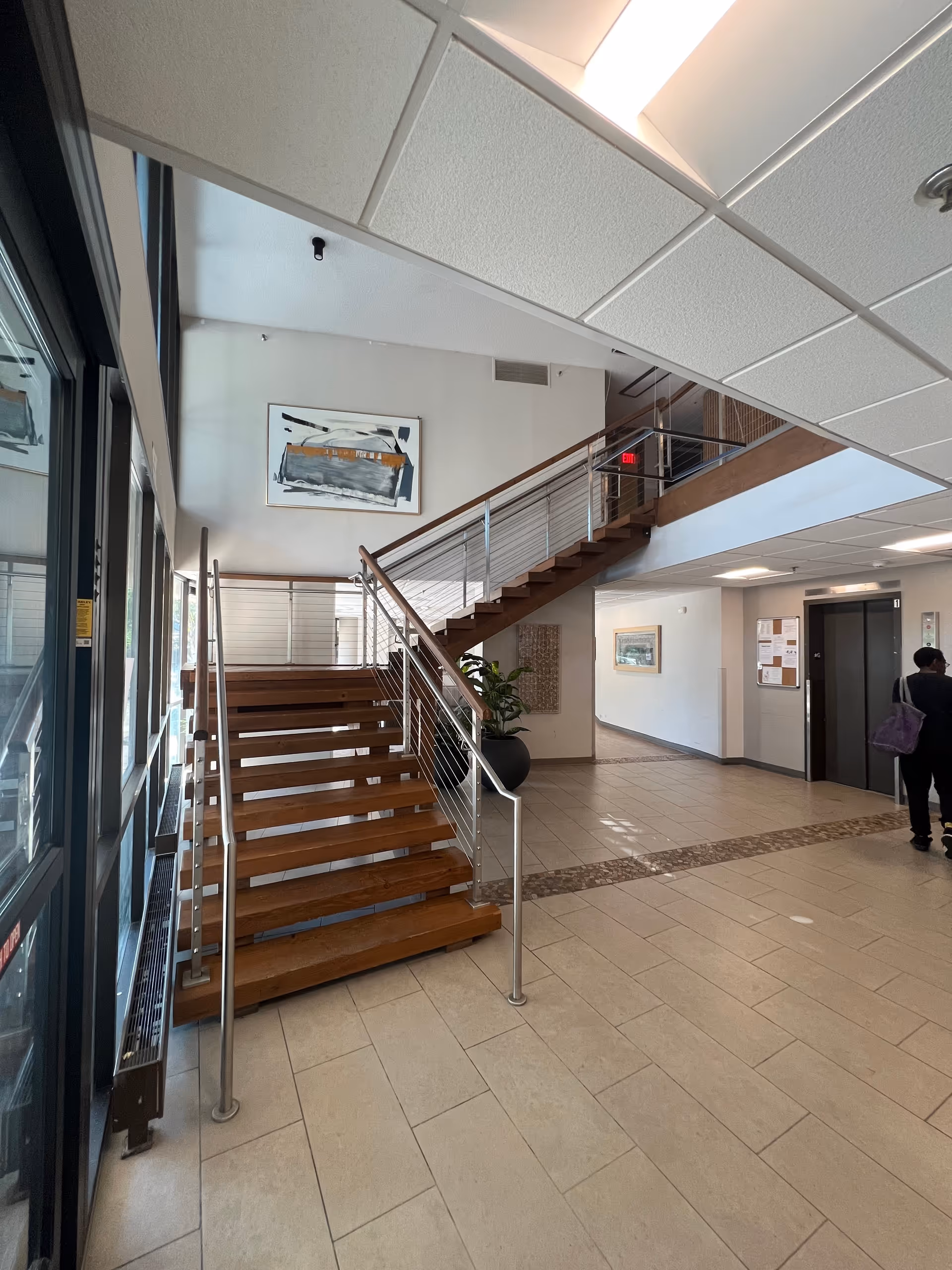 Interior view of a facility lobby featuring a wooden staircase with metal railings leading to an upper floor. The floor is tiled, and there is a large potted plant near the staircase. A person is walking near an elevator on the right side. The walls are decorated with framed artwork and a bulletin board.