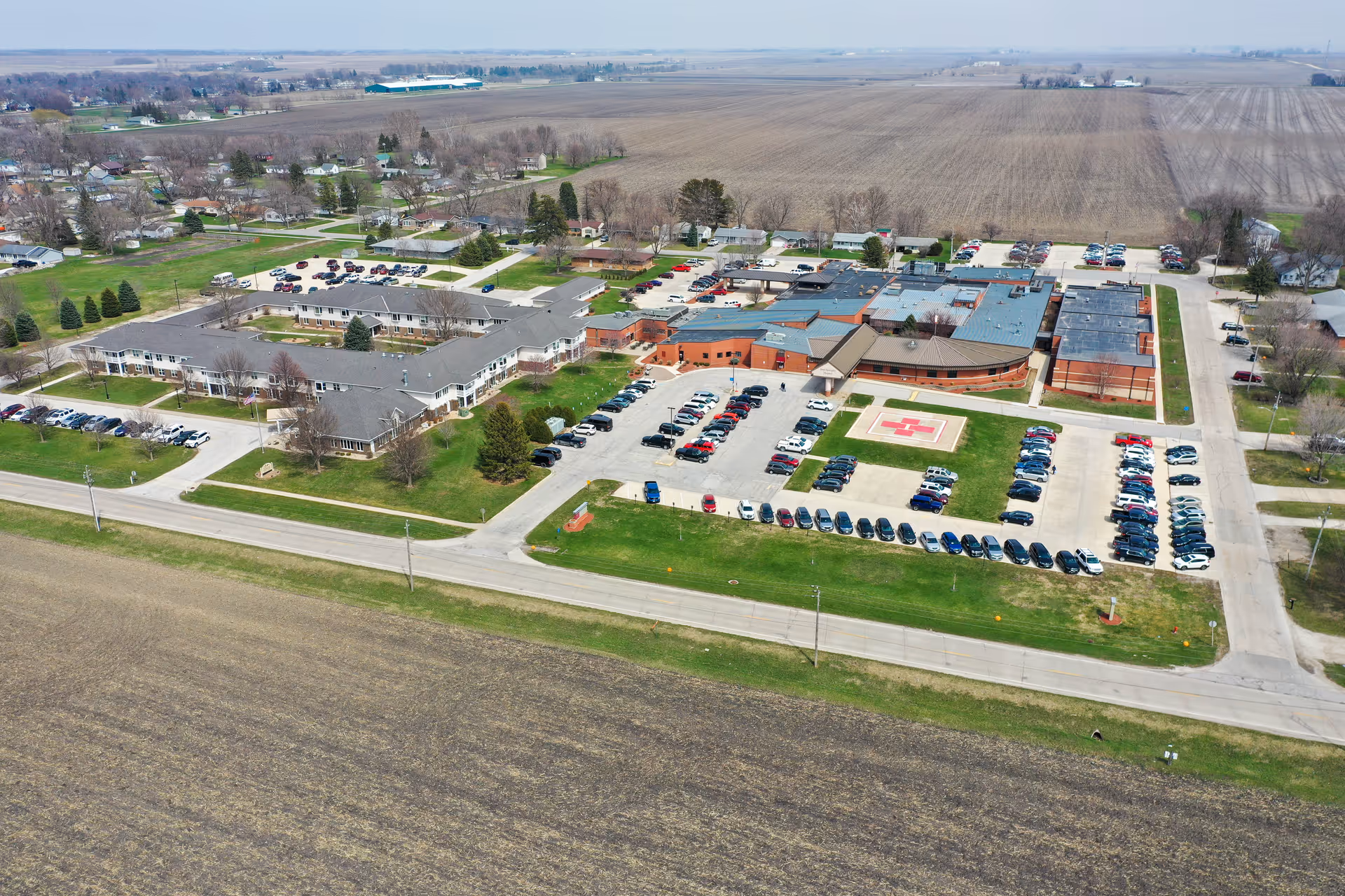 Aerial view of The Meadows Senior Community showing multiple buildings, parking lots filled with cars, green lawns, and surrounding farmland under a clear sky.