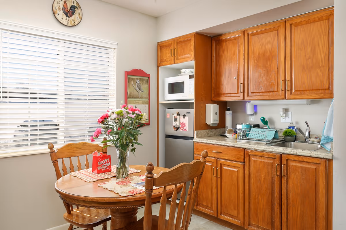 Small kitchenette with wooden cabinets, a round dining table with chairs and a vase of flowers, and a microwave and sink.