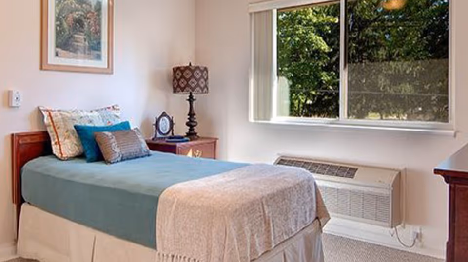 A cozy bedroom with a single bed covered in a blue bedspread and beige blanket. The bed has multiple pillows and is positioned next to a wooden nightstand with a decorative lamp and a small clock. A framed picture hangs on the wall above the bed. A large window lets in natural light and shows greenery outside. Below the window is a wall-mounted air conditioning unit.