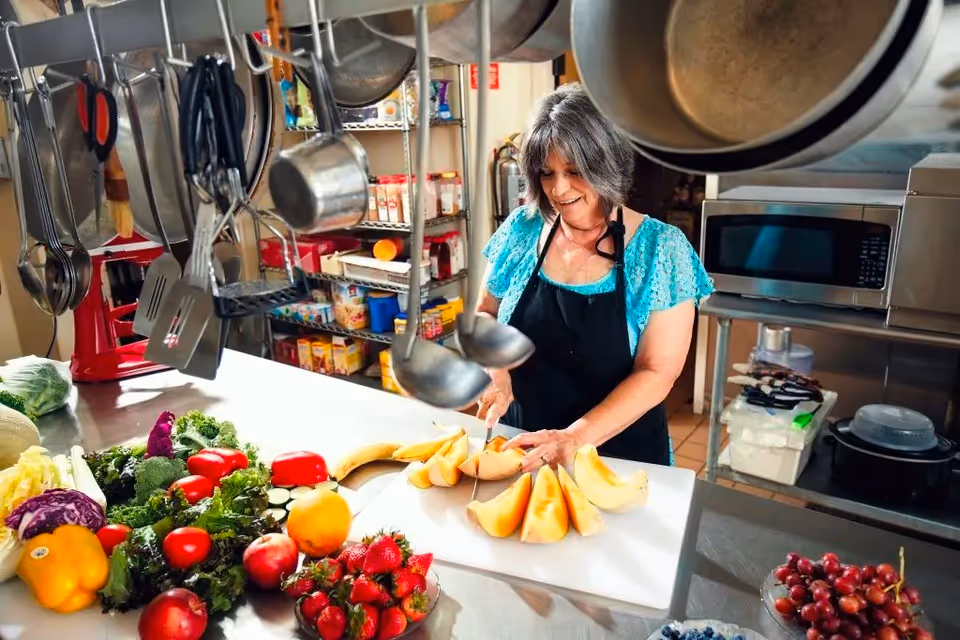 A woman wearing a black apron and turquoise blouse is cutting cantaloupe on a white cutting board in a kitchen. Various fresh fruits and vegetables, including strawberries, grapes, bananas, bell peppers, and leafy greens, are spread out on the counter. Kitchen utensils hang above the counter, and shelves with food items and kitchen appliances are visible in the background.