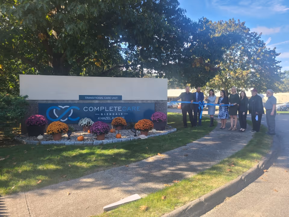 A ribbon-cutting with several people in front of the 'Complete Care Glendale' entrance sign surrounded by flowers and pumpkins.